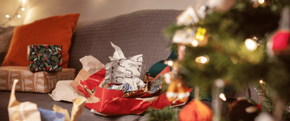 A small pile of festive wrapping paper on a grey sofa. In the foreground, blurred is a Christmas tree with red and gold decorations.