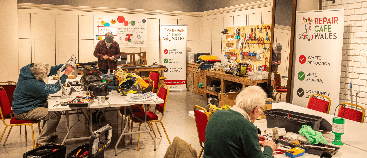 a couple of large table with items ready fo repair. Three people are sat next to the tables, repairing items. Banners that say 'Repair Cafe Wales' are displayed.